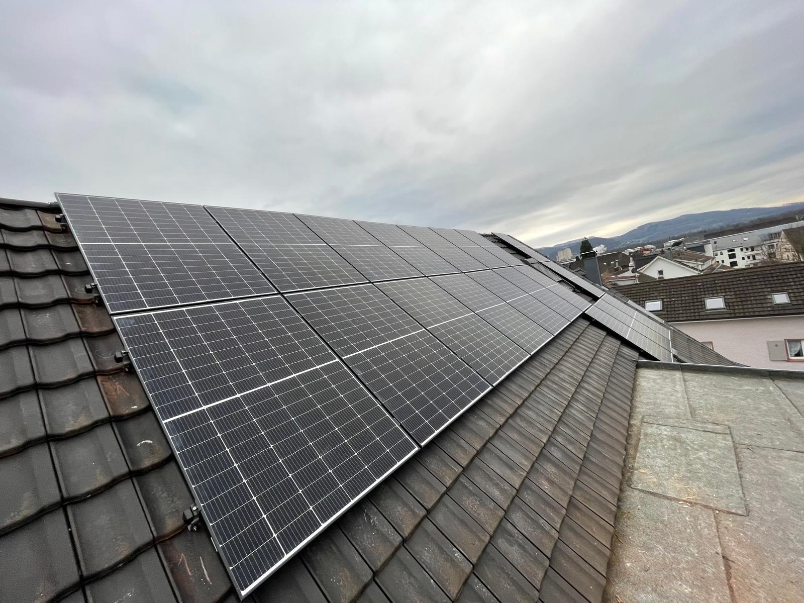 Panele fotowoltaiczne zamontowane na dachu z dachówki ceramicznej, widok z dołu, w tle zabudowa miejska i pochmurne niebo. Instalacja w trakcie dnia.