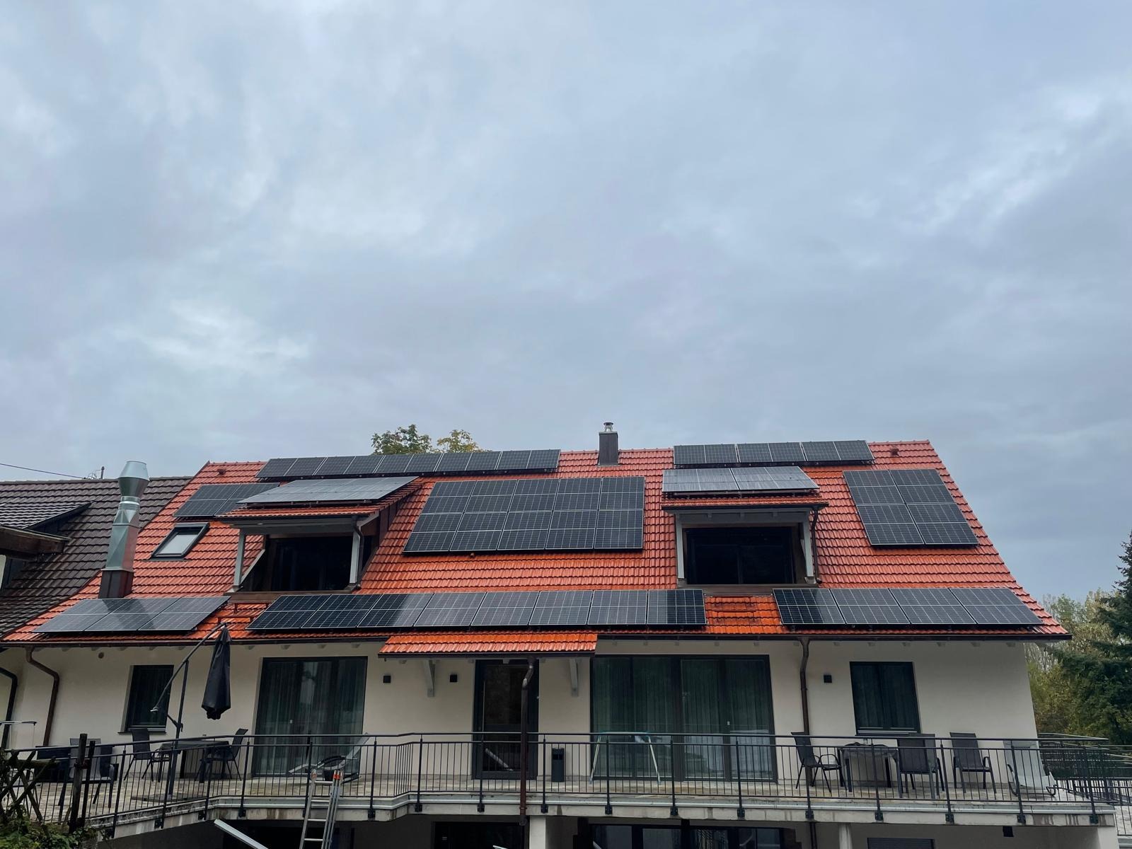 Instalacja paneli słonecznych na dachu domu z czerwoną dachówką. Panele rozmieszczone symetrycznie, widoczne okna dachowe i balkon z metalową balustradą.