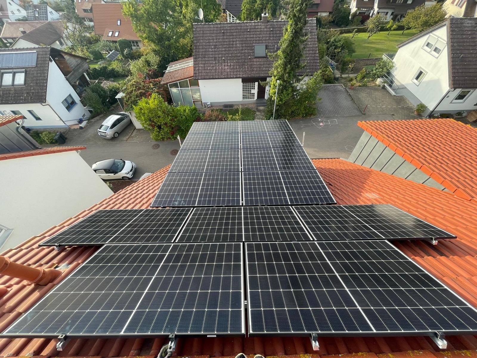 Instalacja paneli fotowoltaicznych na dachu z czerwonej dachówki, widok z góry na otoczenie domów i drzew. Układ paneli w dwóch rzędach, równomierne oświetlenie słoneczne.