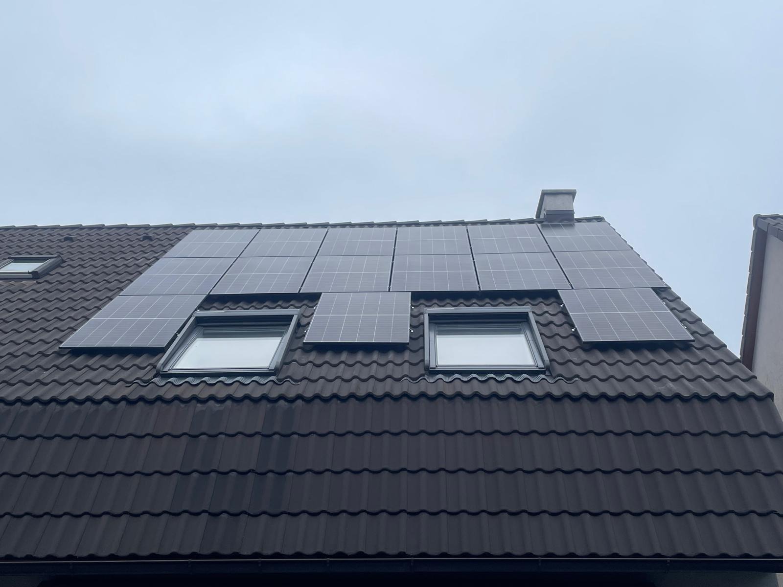 Ciemna dachówka z panelami fotowoltaicznymi wokół okien dachowych. Widok z dołu na zamontowaną instalację w pochmurny dzień.
