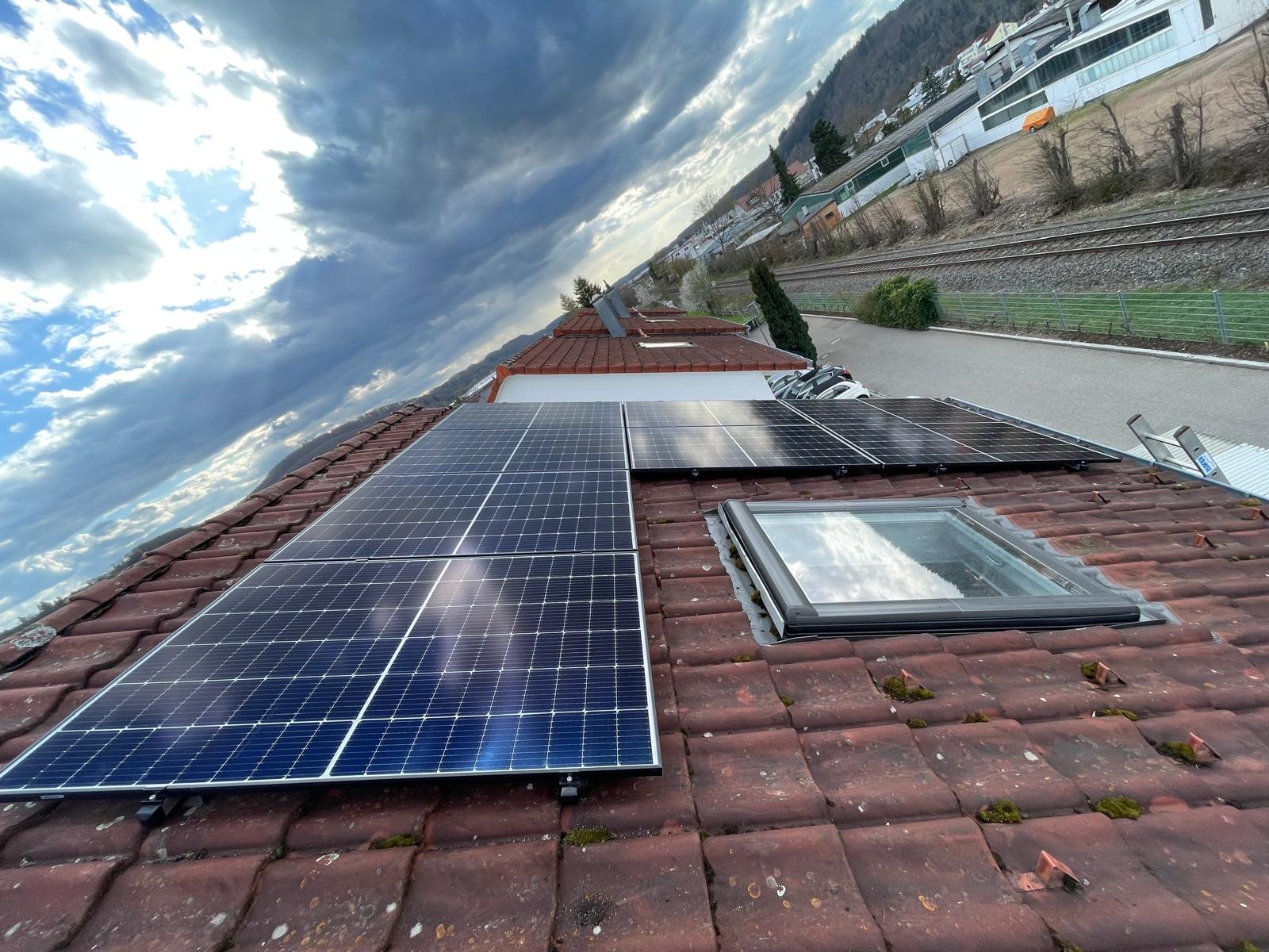 Panele fotowoltaiczne na dachu z czerwoną dachówką, montaż w Tarnowie. Widoczne okno dachowe i drabina. Pochmurne niebo odbija się w panelach.