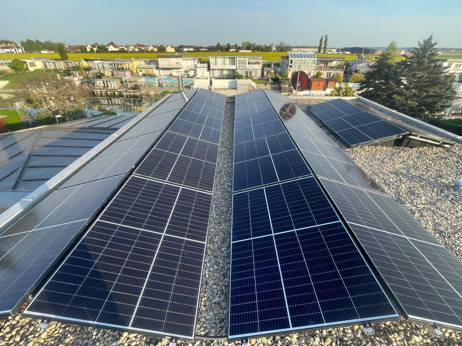 Instalacja fotowoltaiczna na dachu z widokiem na domy i żółte pole. Panele ułożone rzędami, oddzielone żwirem. Słoneczny dzień, błękitne niebo.