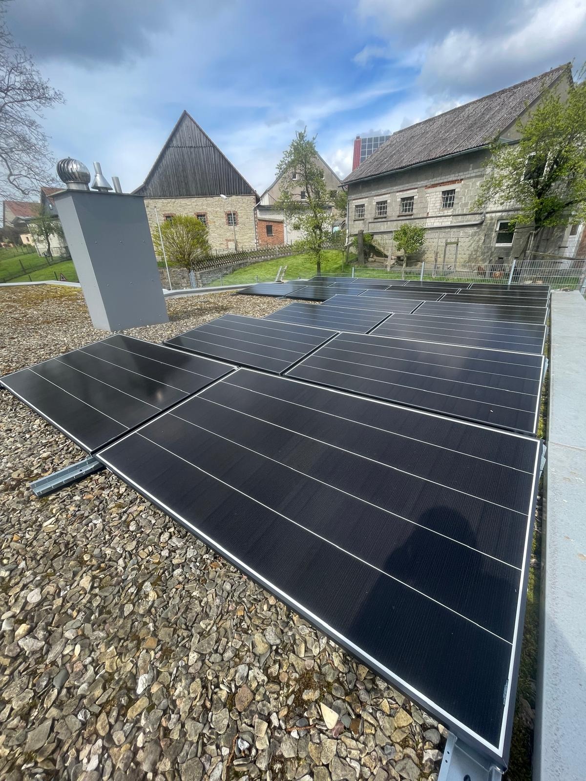 Instalacja paneli fotowoltaicznych na dachu budynku z kamieniami, z widocznym systemem wentylacji i zabudową w tle. Ujęcie z bliska.