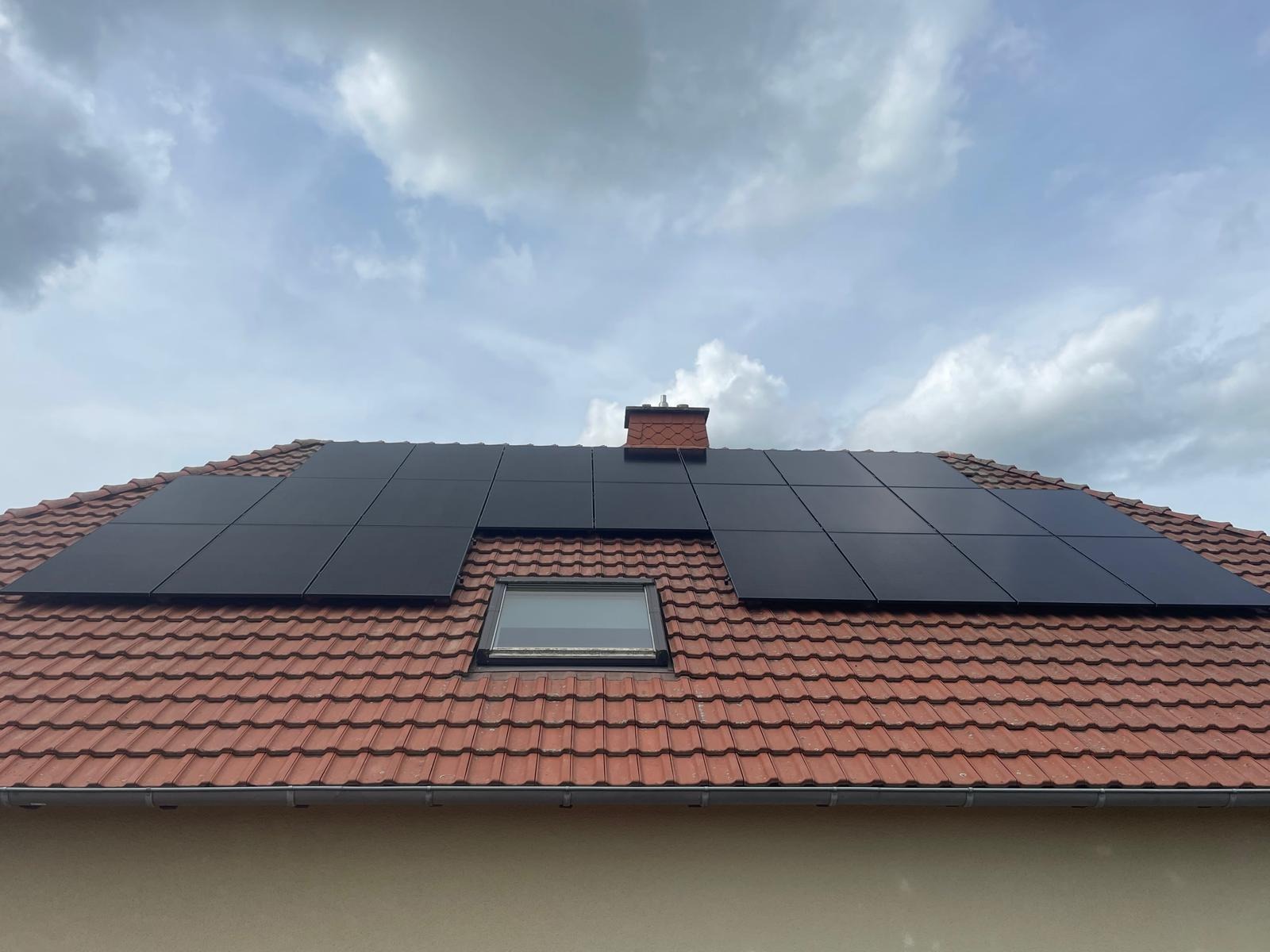 Czerwony dach z panelami fotowoltaicznymi w układzie poziomym i oknem dachowym, widok z dołu, na tle pochmurnego nieba.