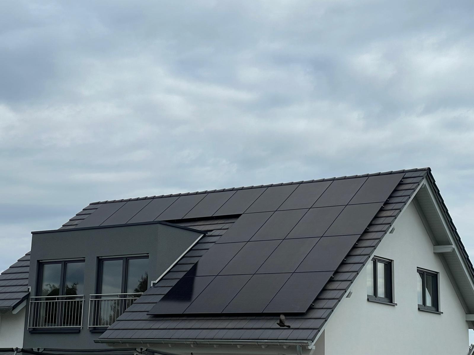 Ciemne panele fotowoltaiczne na dachu nowoczesnego domu z balkonem, widoczne na tle pochmurnego nieba. Minimalistyczny design paneli.