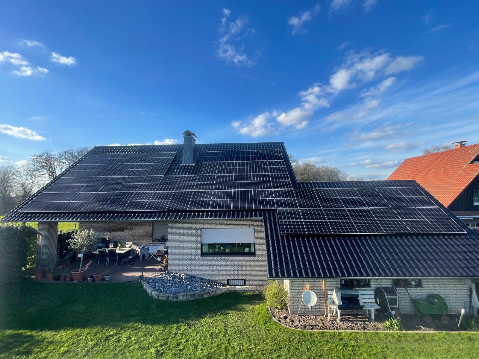 Dom jednorodzinny z panelami fotowoltaicznymi na dachu, widok z ogrodu w słoneczny dzień. Biała cegła, czarna dachówka i błękitne niebo z delikatnymi chmurami.