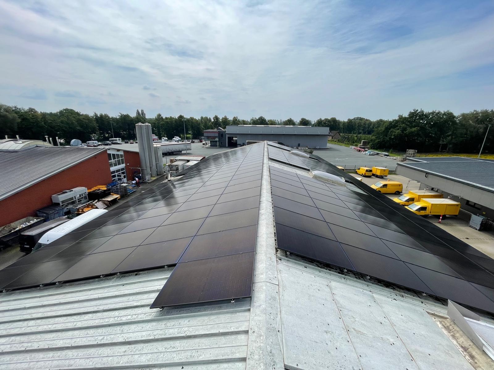 Instalacja paneli fotowoltaicznych na dachu budynku przemysłowego, widok z góry, żółte samochody dostawcze w tle, pochmurne niebo.