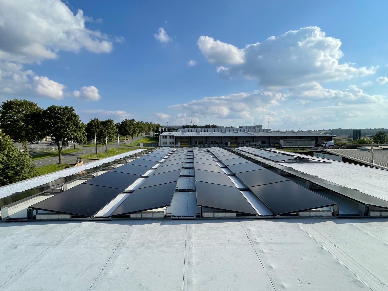 Równomiernie rozłożone panele fotowoltaiczne na dachu budynku przemysłowego w słoneczny dzień, widok z góry, błękitne niebo z chmurami w tle.