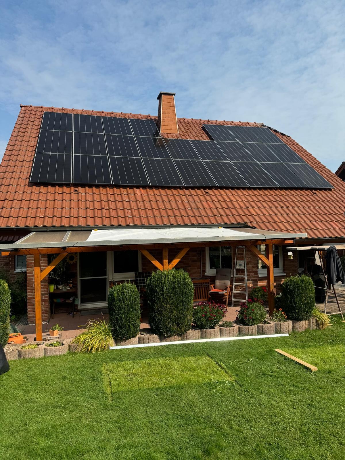 Instalacja paneli słonecznych na dachu domu z czerwoną dachówką. Widoczny ogród i pergola. Drabina oparta o ścianę. Słoneczny dzień.