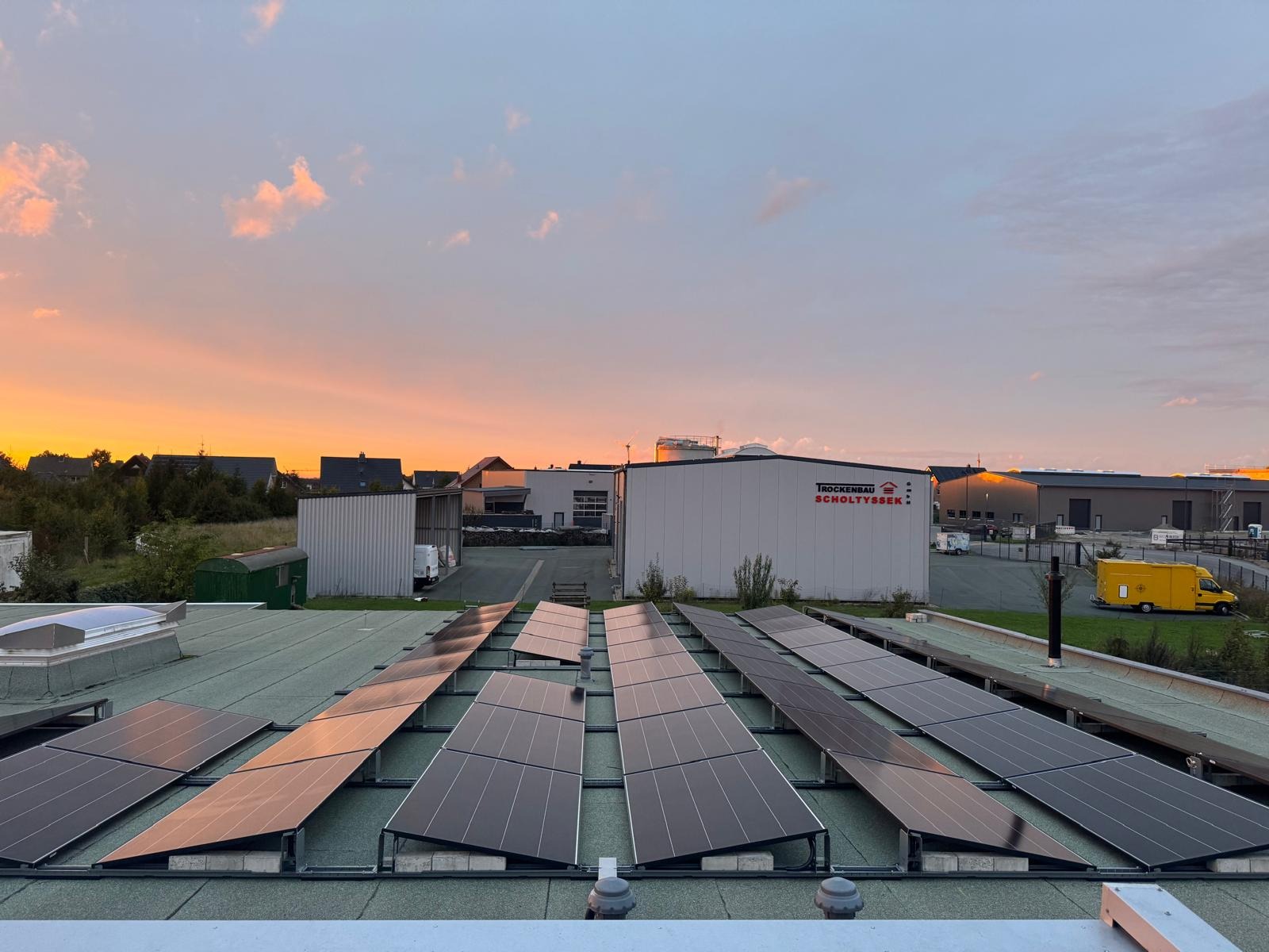 Panele fotowoltaiczne na dachu budynku o zmierzchu, odbijające pomarańczowe światło. W tle zabudowania przemysłowe i fragmenty zieleni. Widok z góry.