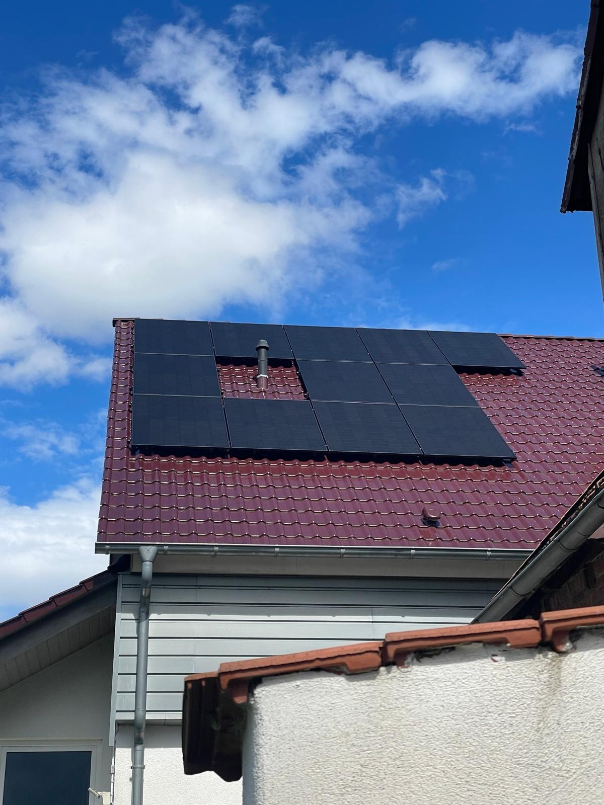 Panele fotowoltaiczne na dachu domu z czerwoną dachówką, widoczne na tle błękitnego nieba z chmurami. Kominek wystaje z dachu obok paneli.