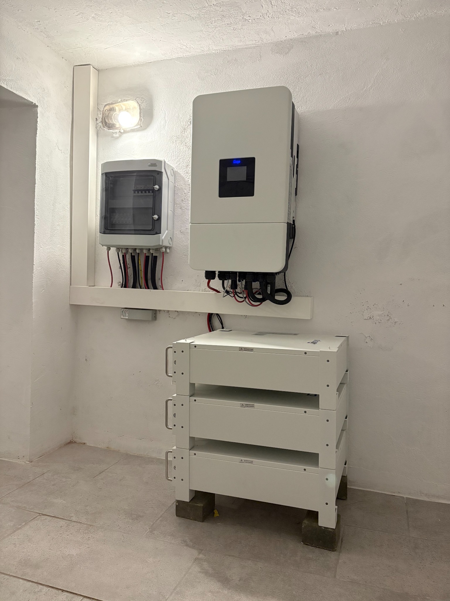 Kompletna instalacja magazynu energii: trzy moduły bateryjne na betonowych podstawach, inwerter i rozdzielnica na ścianie w jasnym pomieszczeniu.