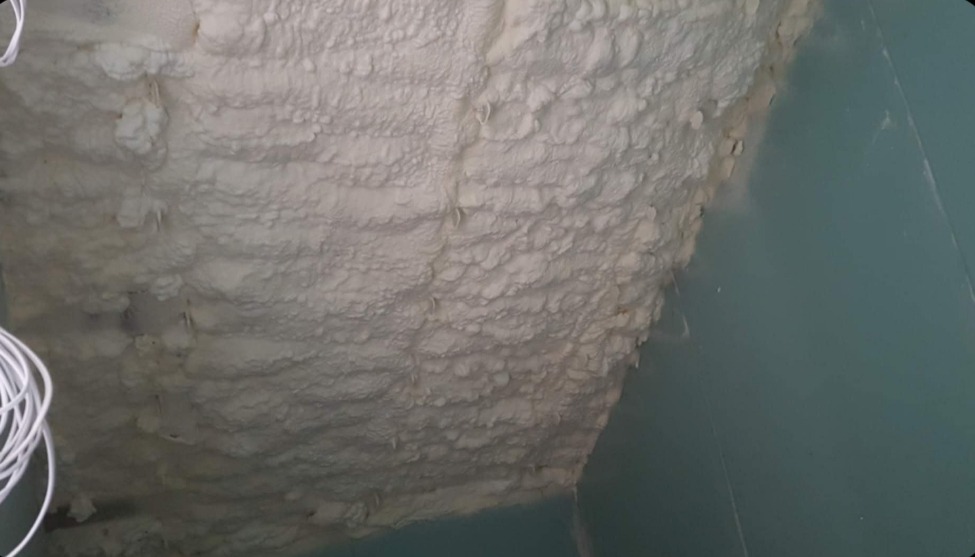 Ściana pokryta grubą warstwą białej pianki izolacyjnej PUR, widoczne nierówności i struktura piany. Fragment instalacji elektrycznej w lewym dolnym rogu.