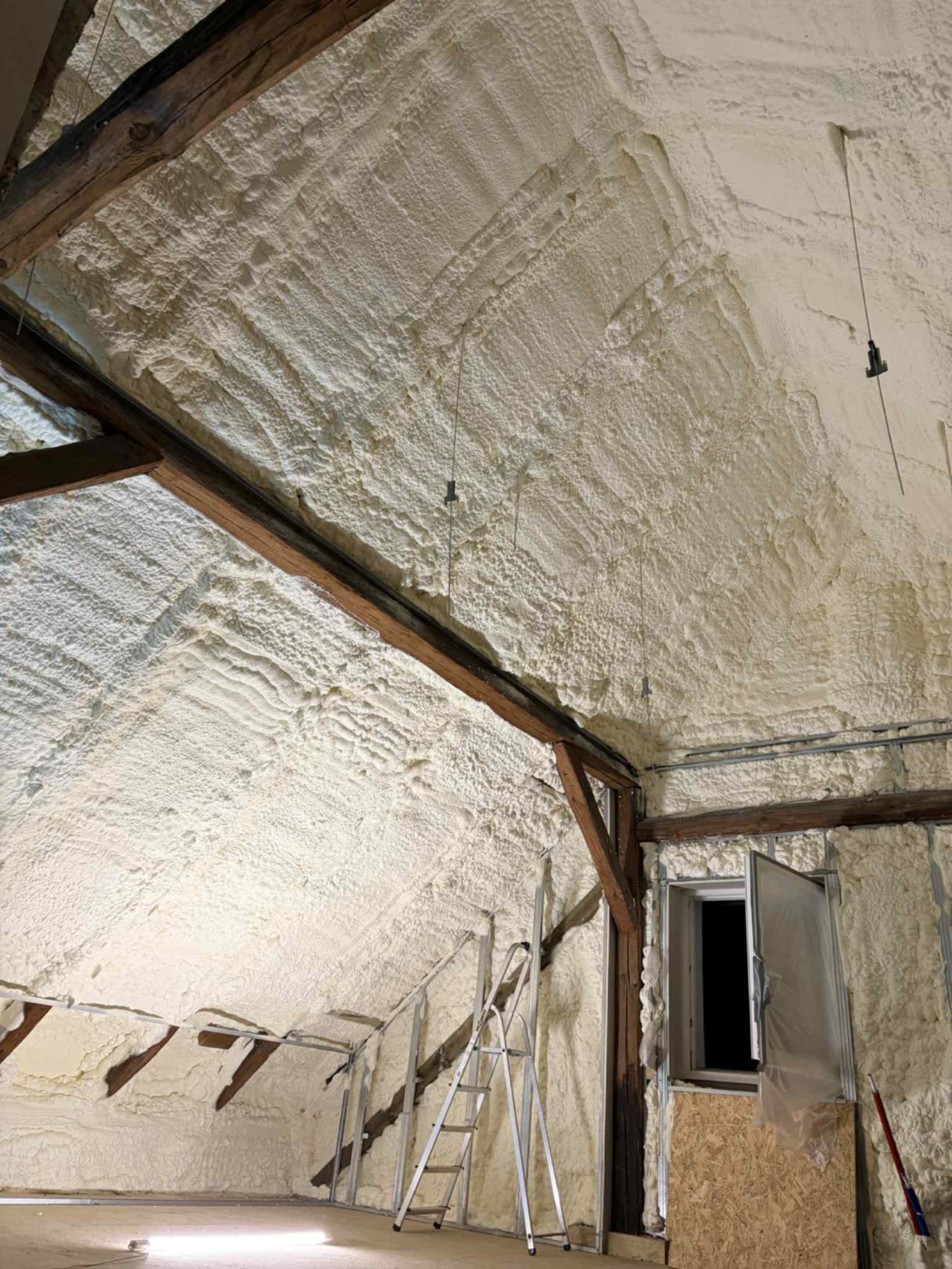 Poddasze po ociepleniu pianką PUR, widoczne drewniane belki i okno. Biała, nierówna powierzchnia pianki pokrywa ściany i sufit. Metalowa drabina oparta o ścianę.