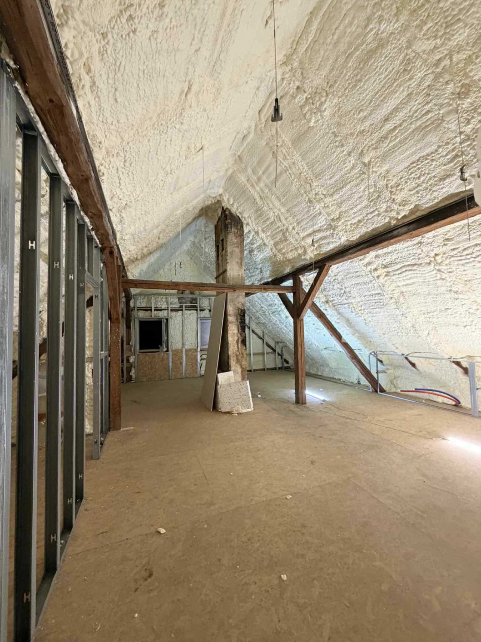 Poddasze z drewnianą konstrukcją i metalowymi profilami, pokryte grubą warstwą białej pianki izolacyjnej na skośnym dachu. Widoczny komin i fragment instalacji.
