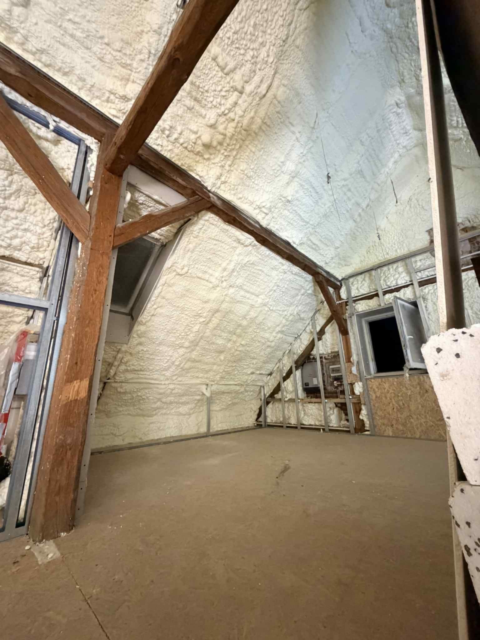 Poddasze po ociepleniu pianką PUR: widoczne drewniane belki konstrukcyjne i okna dachowe. Biała pianka pokrywa skośne ściany i sufit, tworząc jednolitą warstwę izolacyjną.