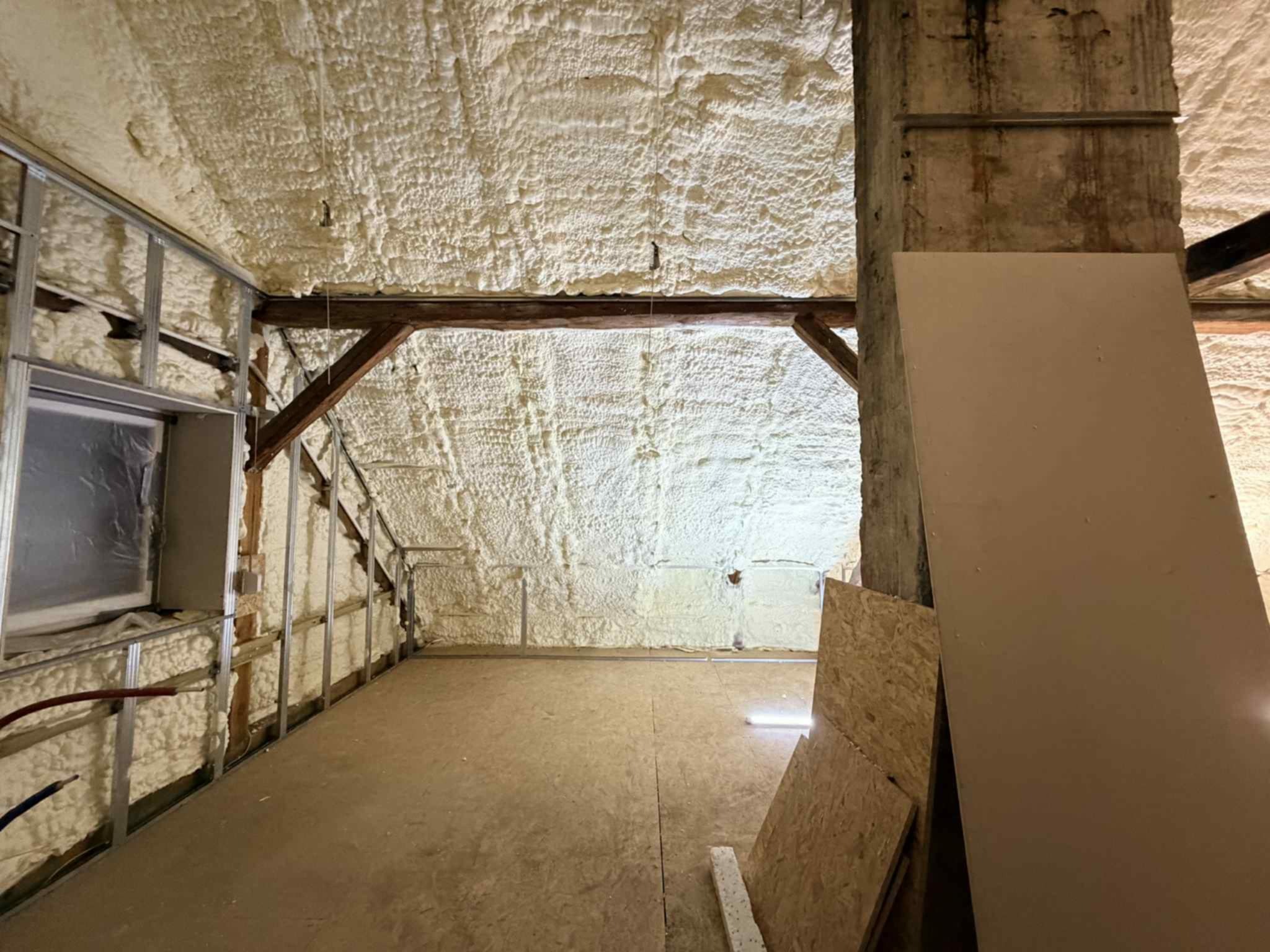 Poddasze po ociepleniu pianką PUR, widoczne drewniane belki, metalowy stelaż i płyty OSB. Biała pianka pokrywa ściany i sufit, tworząc jednolitą warstwę izolacyjną.