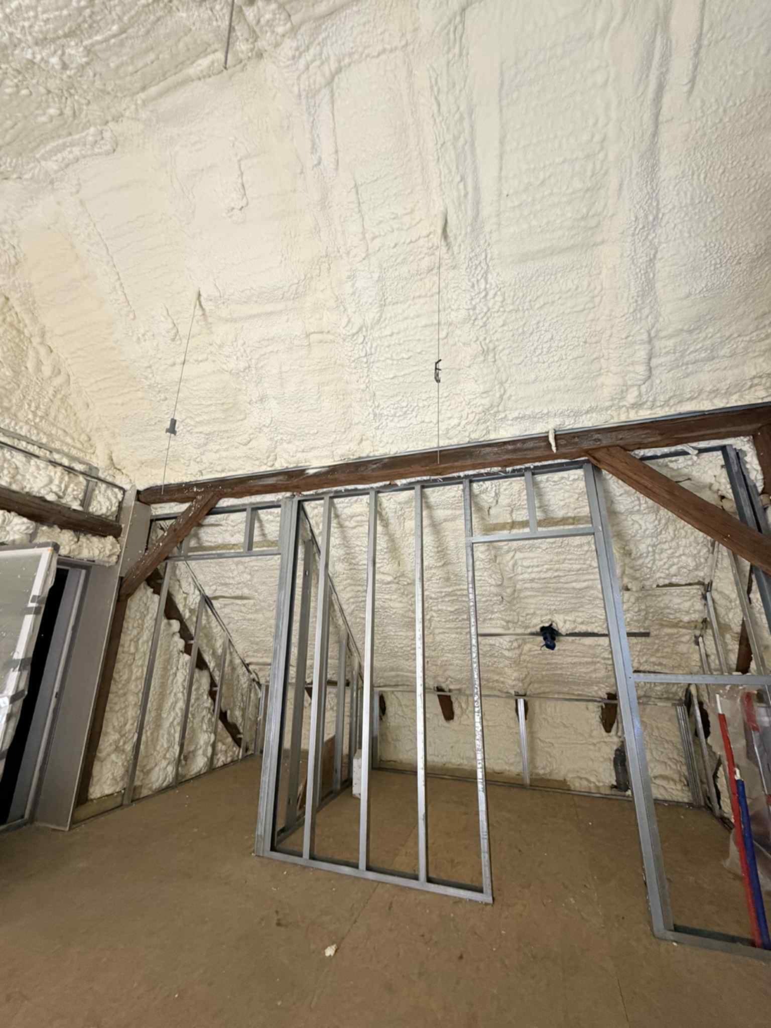 Poddasze z widoczną konstrukcją szkieletową i grubą warstwą białej pianki izolacyjnej na skośnym suficie i ścianach, ukazujące etap prac ociepleniowych.