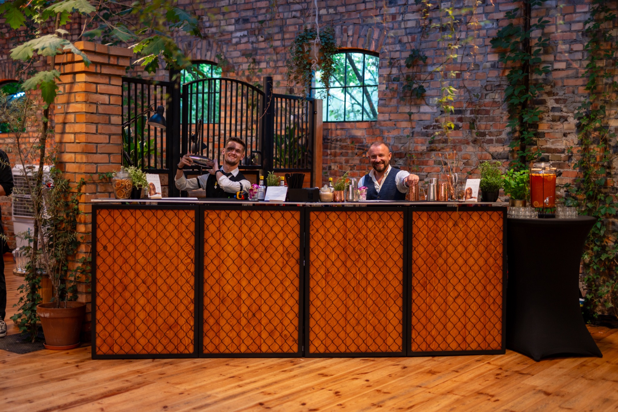Barmani za drewnianym barem z kratką na tle ceglanej ściany z bluszczem i oknami. Jeden barman potrząsa shakerem, drugi uśmiecha się, wskazując gestem na widza.