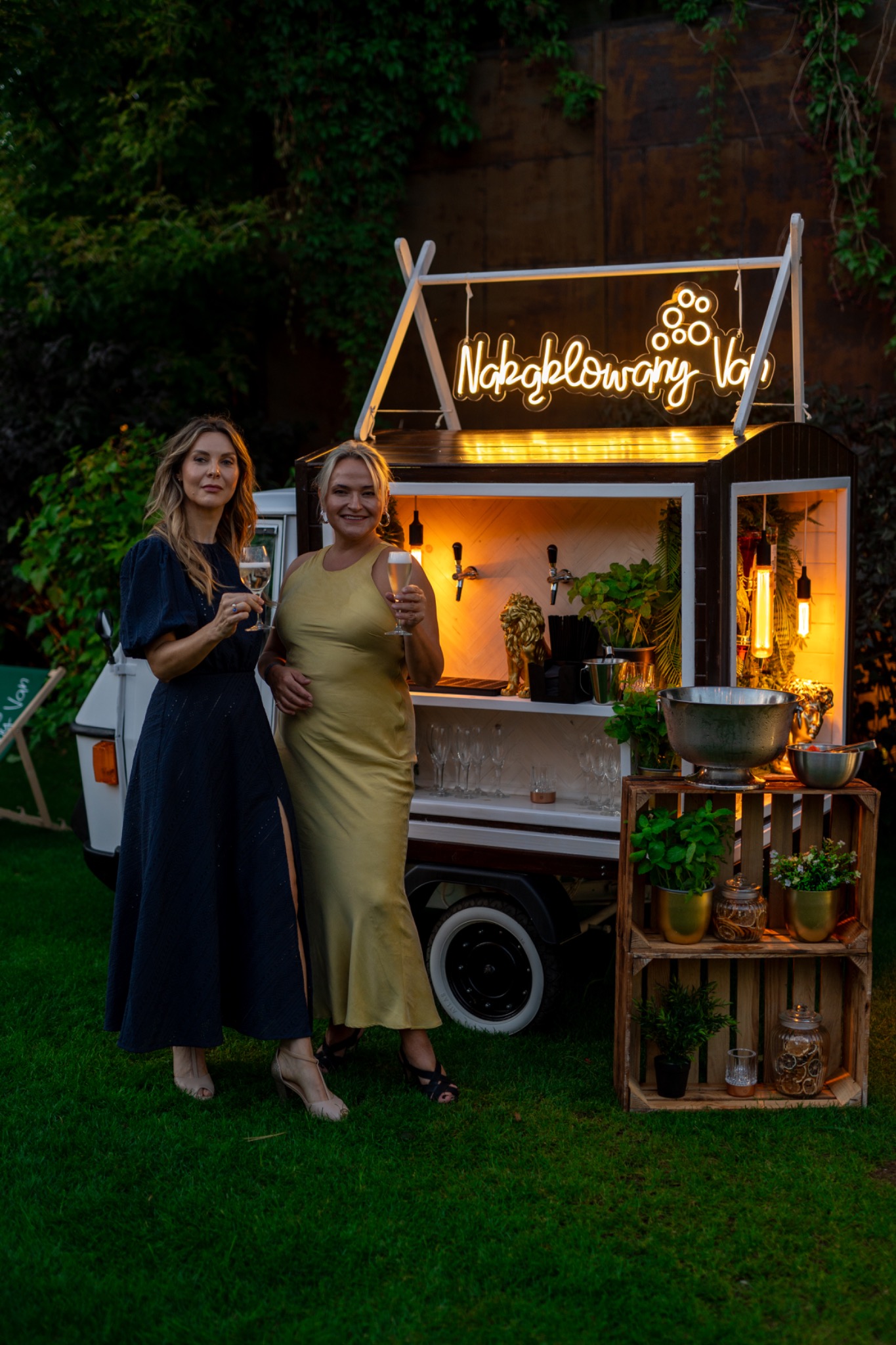 Dwie kobiety z kieliszkami szampana stoją przed mobilnym barem z neonowym napisem 'Nakręcony Van' w ogrodzie. Bar wyposażony w kraniki i dekoracje roślinne.