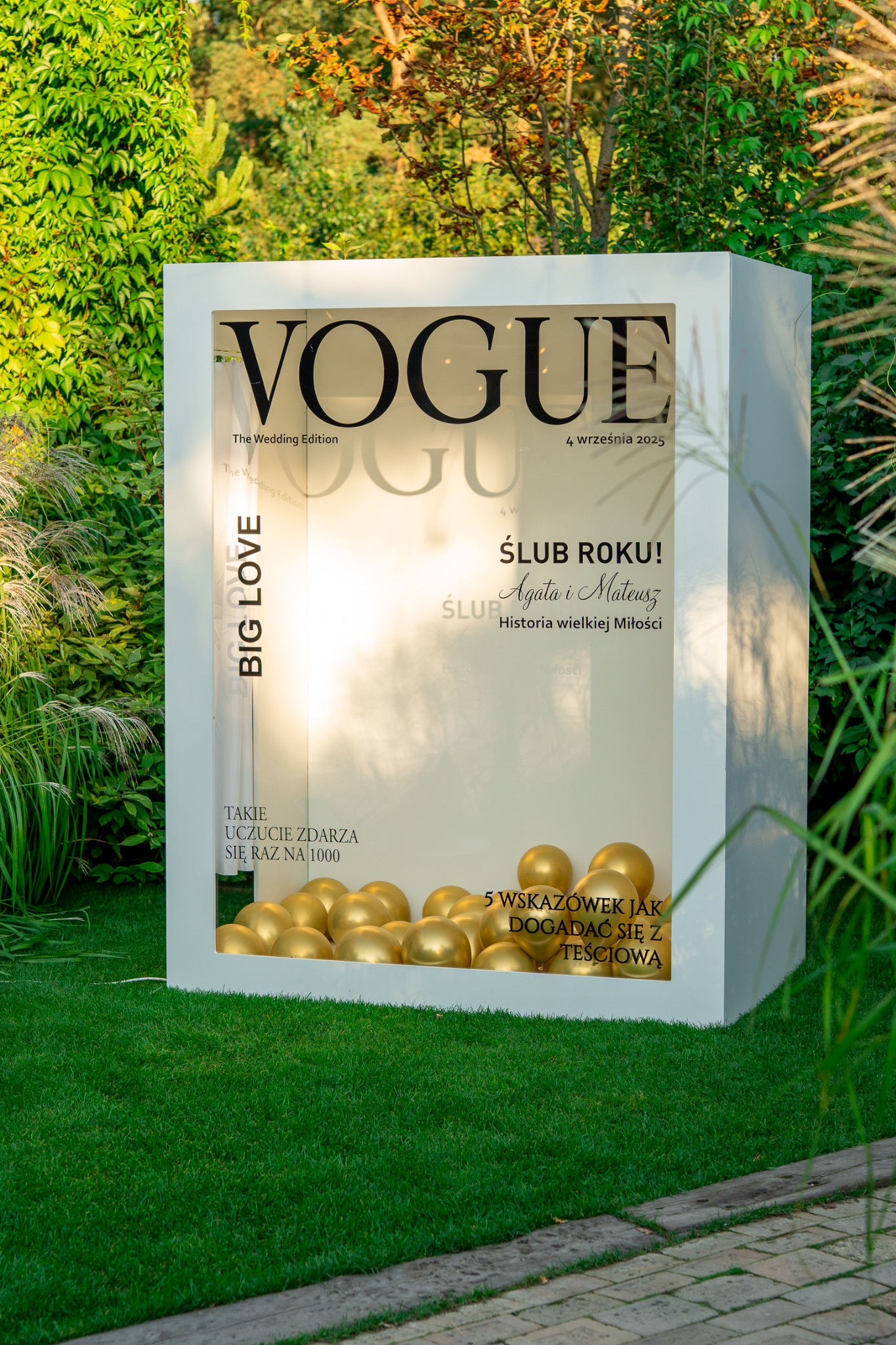 Dekoracja ślubna w plenerze imitująca okładkę magazynu Vogue z napisem 'Ślub Roku!' i złotymi balonami, otoczona zielenią.
