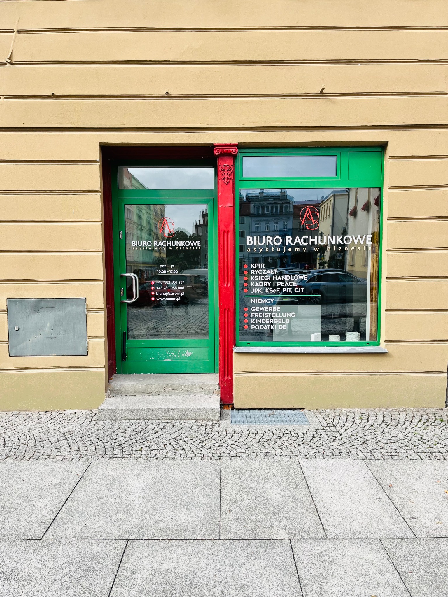 Fasada biura rachunkowego z zielonymi drzwiami i oknami, widoczny napis 'Biuro Rachunkowe asystujemy w biznesie' oraz zakres usług. Budynek w odcieniach beżu.