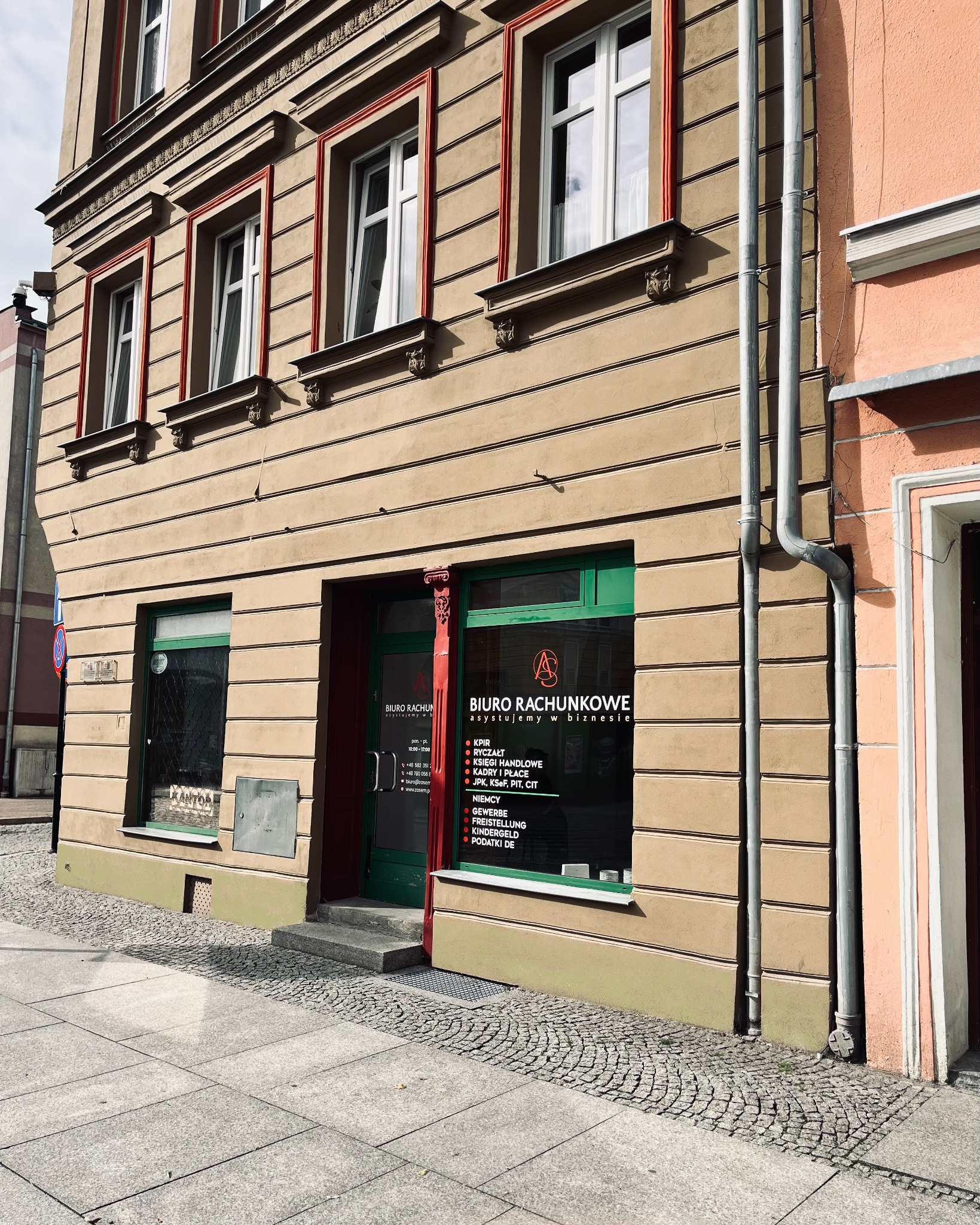 Fasada biura rachunkowego w kamienicy z zielonymi ramami okiennymi i czerwonymi elementami dekoracyjnymi. Na oknie widoczne informacje o usługach.