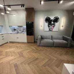 Nowoczesne wnętrze: jasna kuchnia z marmurowym blatem, drewniana podłoga w jodełkę, szara sofa i grafika na ścianie. Oświetlenie punktowe i designerska lampa.