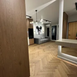 Nowoczesne wnętrze: drewniana podłoga w jodełkę, szara sofa i abstrakcyjny obraz. Widoczna zabudowa kuchenna z drewnianymi frontami i oświetlenie punktowe.