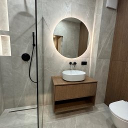 WroTeam Meble sp. z o.o. - Nowoczesna łazienka z kabiną prysznicową typu walk-in, okrągłym lustrem z podświetleniem LED i szafką z umywalką w odcieniach drewna, wykończona płytkami imitującymi marmur.