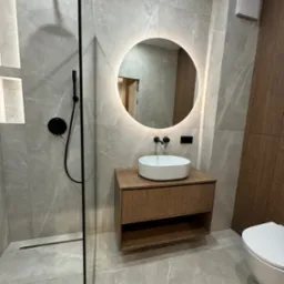 Nowoczesna łazienka z kabiną prysznicową typu walk-in, okrągłym lustrem z podświetleniem LED i szafką z umywalką w odcieniach drewna, wykończona płytkami imitującymi marmur.