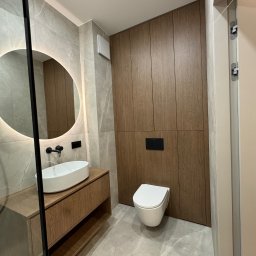 WroTeam Meble sp. z o.o. - Nowoczesna łazienka z drewnianą zabudową, okrągłym lustrem z podświetleniem LED i wiszącą toaletą. Minimalistyczny design, płytki imitujące marmur.