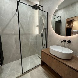 WroTeam Meble sp. z o.o. - Nowoczesna łazienka z kabiną typu walk-in, drewnianą szafką pod umywalkę i okrągłym lustrem z podświetleniem LED. Szare płytki imitujące marmur na ścianach i podłodze.