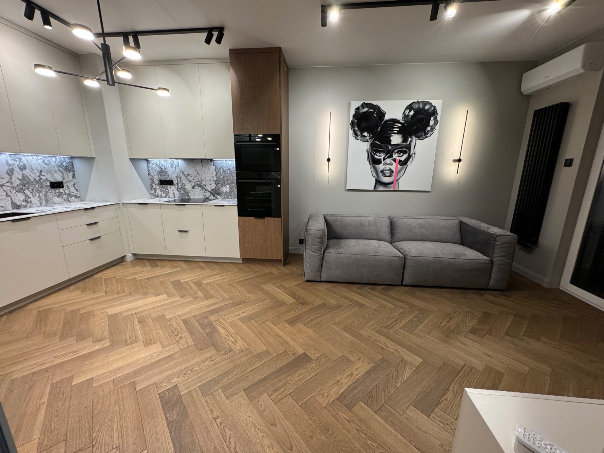Nowoczesne wnętrze: jasna kuchnia z marmurowym blatem, drewniana podłoga w jodełkę, szara sofa i grafika na ścianie. Oświetlenie punktowe i designerska lampa.