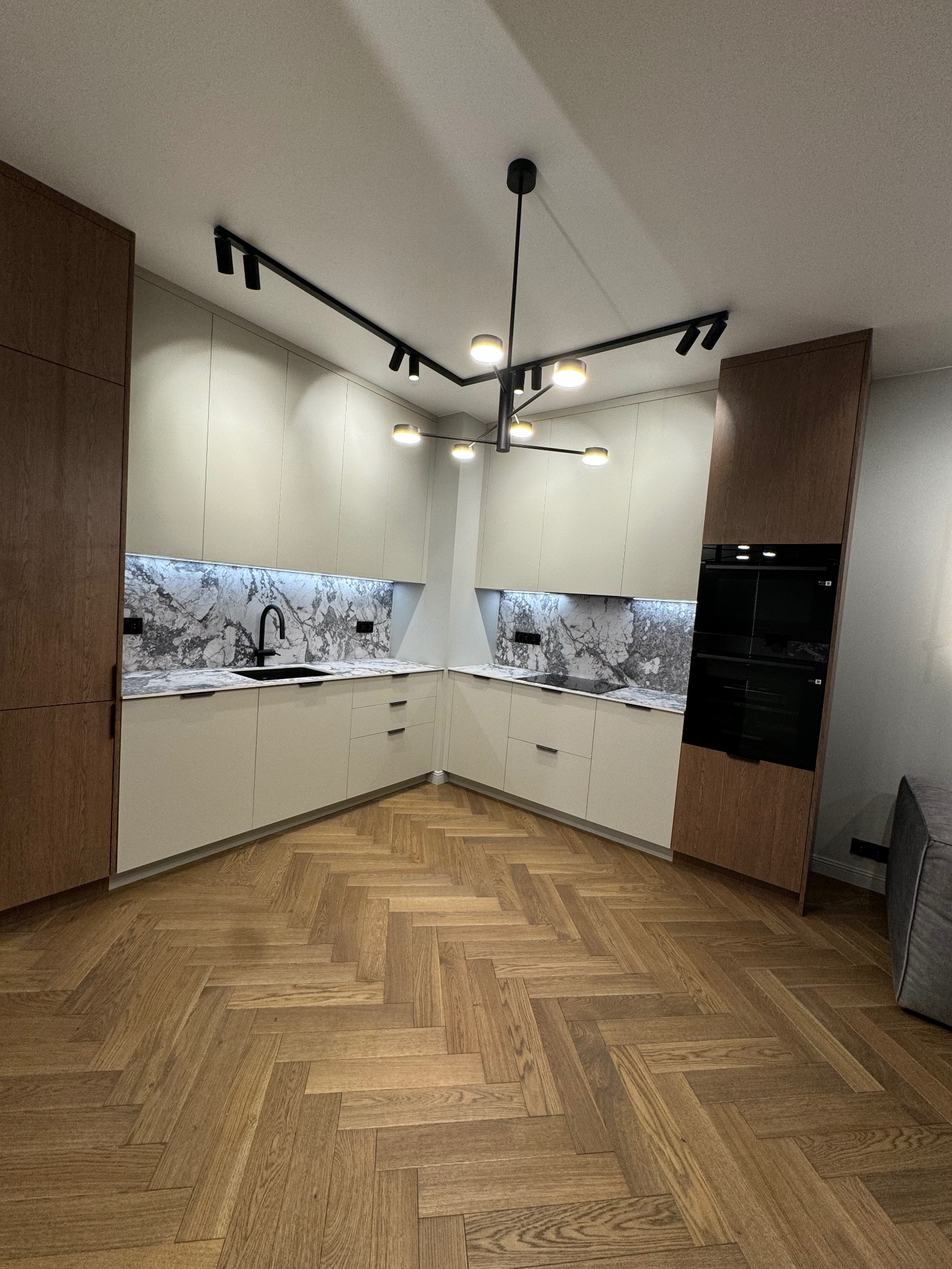 Nowoczesna kuchnia w kształcie litery U z drewnianą podłogą w jodełkę, kremowymi szafkami i marmurowym backsplash. Czarna armatura i oświetlenie punktowe dopełniają całość.