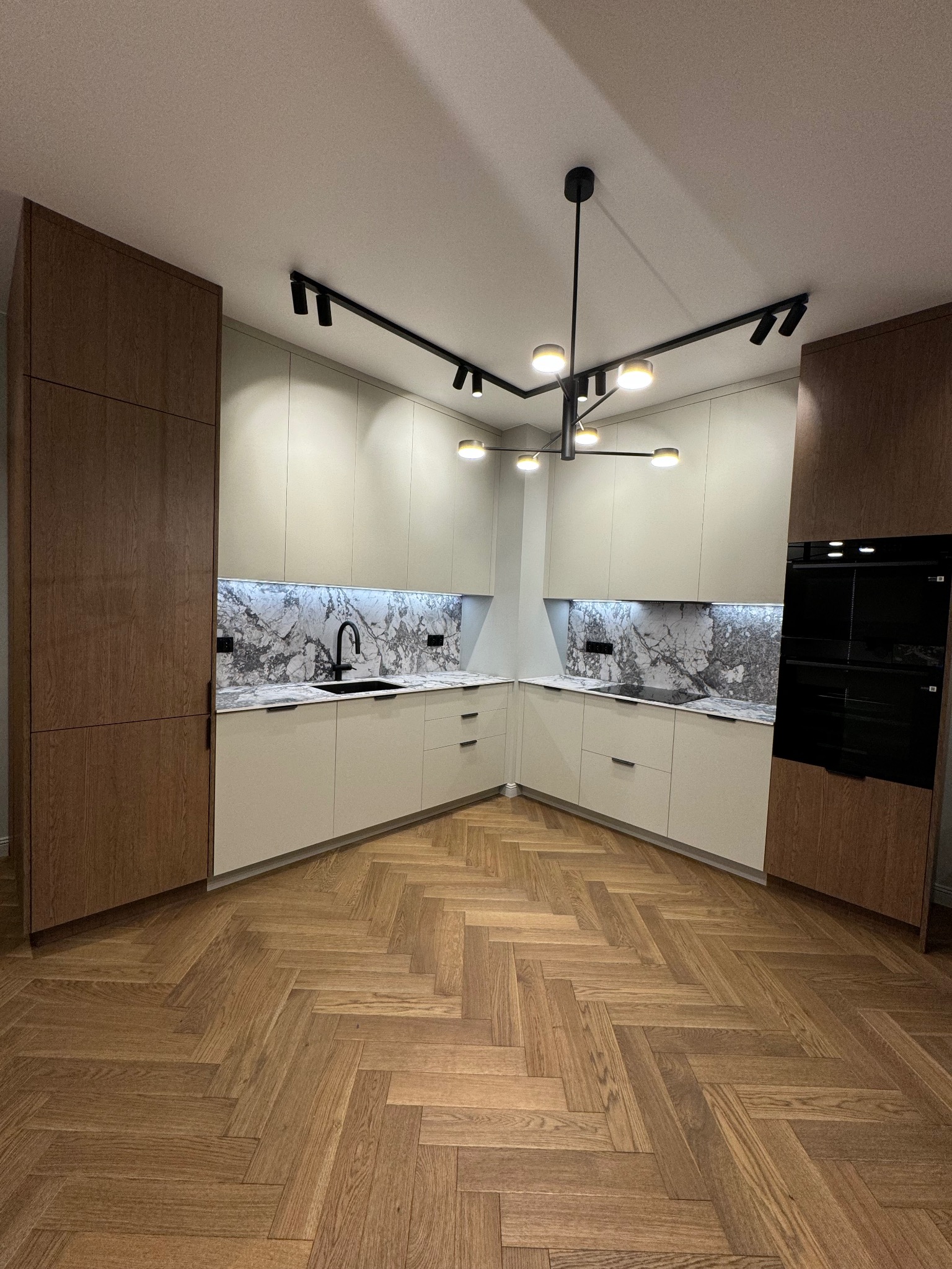 Nowoczesna kuchnia w kształcie litery U z drewnianą podłogą w jodełkę. Kremowe szafki, marmurowy backsplash i czarna armatura. Oświetlenie punktowe i designerska lampa.