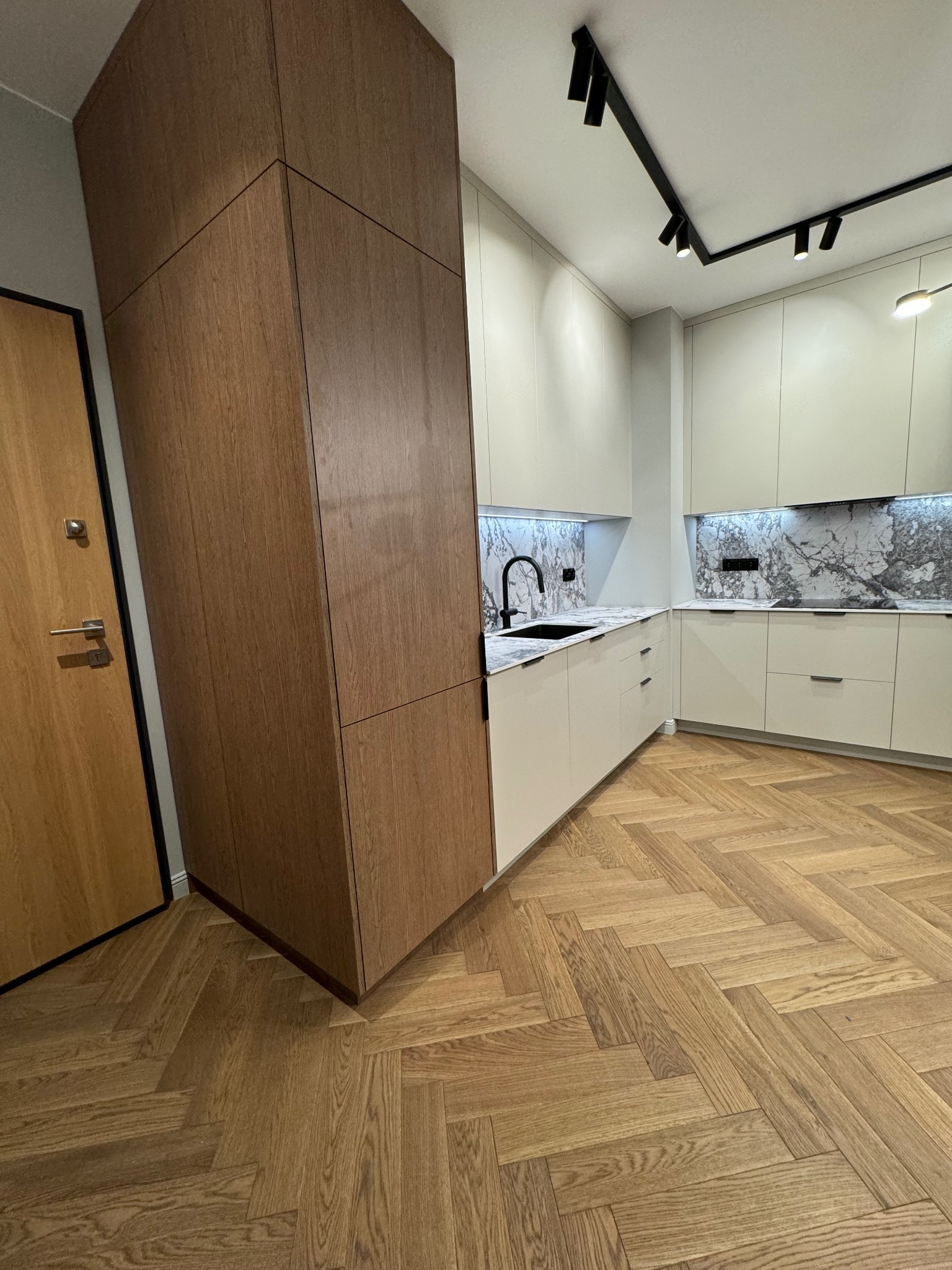 Nowoczesna kuchnia: jasne szafki z marmurowym backsplash'em, drewniana kolumna szaf, parkiet w jodełkę i oświetlenie punktowe. Minimalistyczny design.