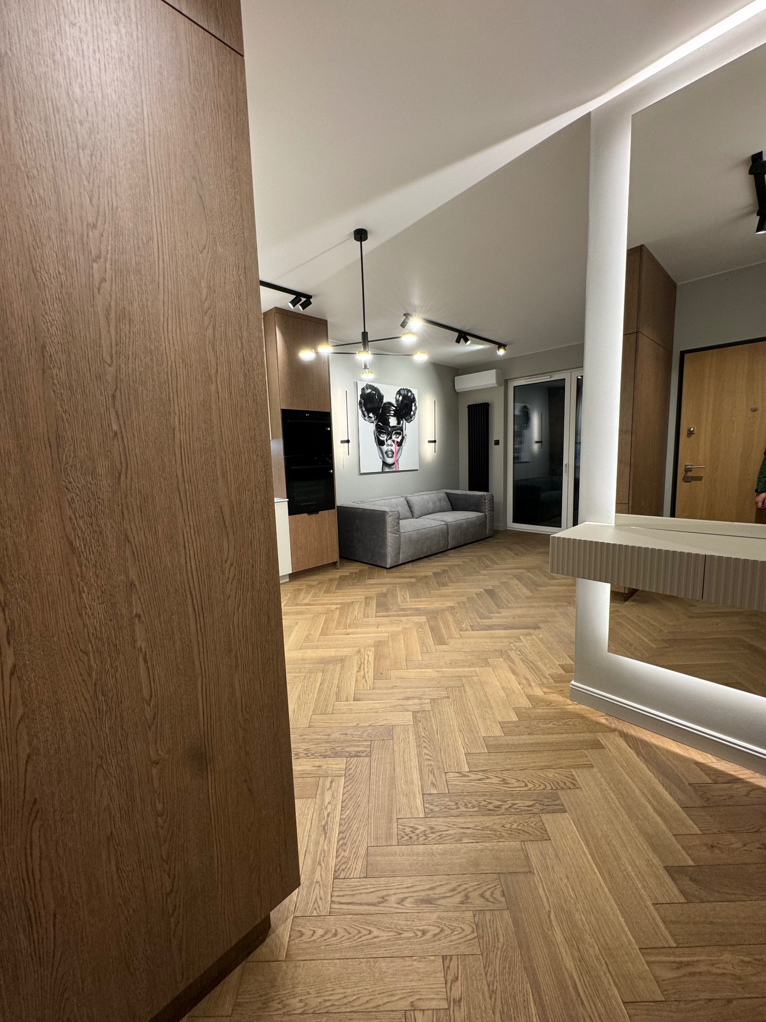 Nowoczesne wnętrze: drewniana podłoga w jodełkę, szara sofa i abstrakcyjny obraz. Widoczna zabudowa kuchenna z drewnianymi frontami i oświetlenie punktowe.