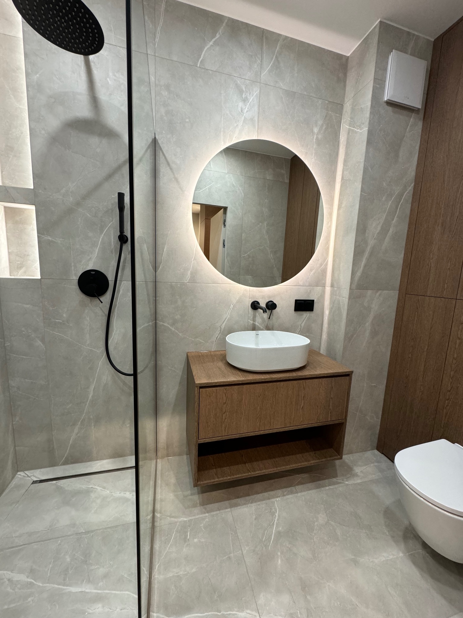 Nowoczesna łazienka z kabiną prysznicową typu walk-in, okrągłym lustrem z podświetleniem LED i szafką z umywalką w odcieniach drewna, wykończona płytkami imitującymi marmur.