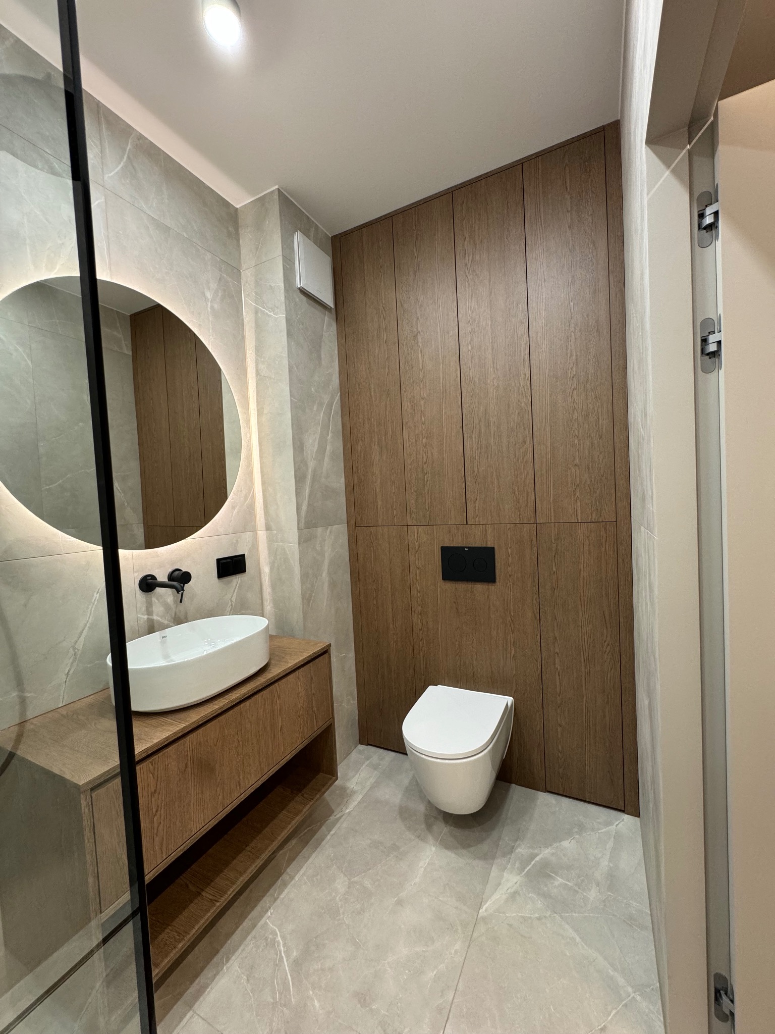 Nowoczesna łazienka z drewnianą zabudową, okrągłym lustrem z podświetleniem LED i wiszącą toaletą. Minimalistyczny design, płytki imitujące marmur.
