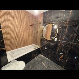 Renew Home sp.z.o.o - Elegancka łazienka z czarnymi płytkami imitującymi marmur, drewnianą ścianą, okrągłym lustrem w złotej ramie i kabiną prysznicową ze złotymi elementami.