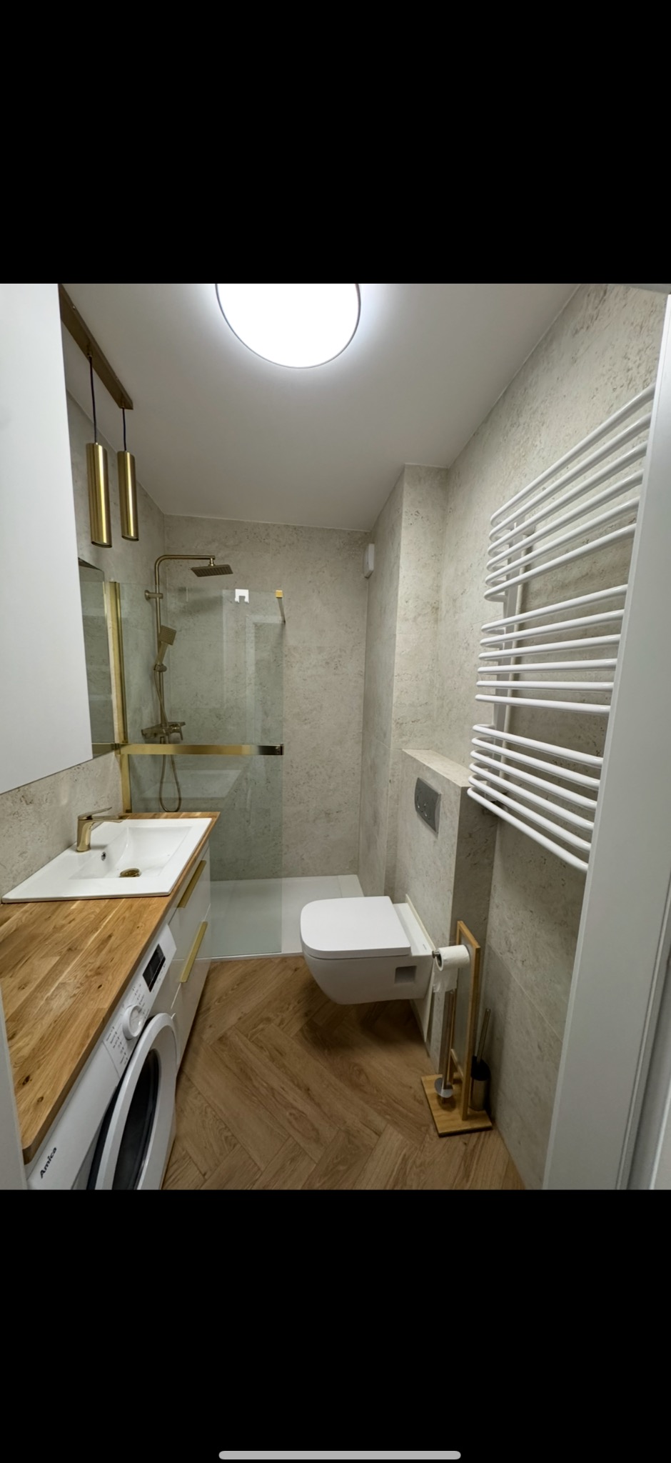 Nowoczesna łazienka z drewnianym blatem i złotymi akcentami. Umywalka w zabudowie z pralką, kabina prysznicowa typu walk-in, grzejnik drabinkowy i parkiet w jodełkę.