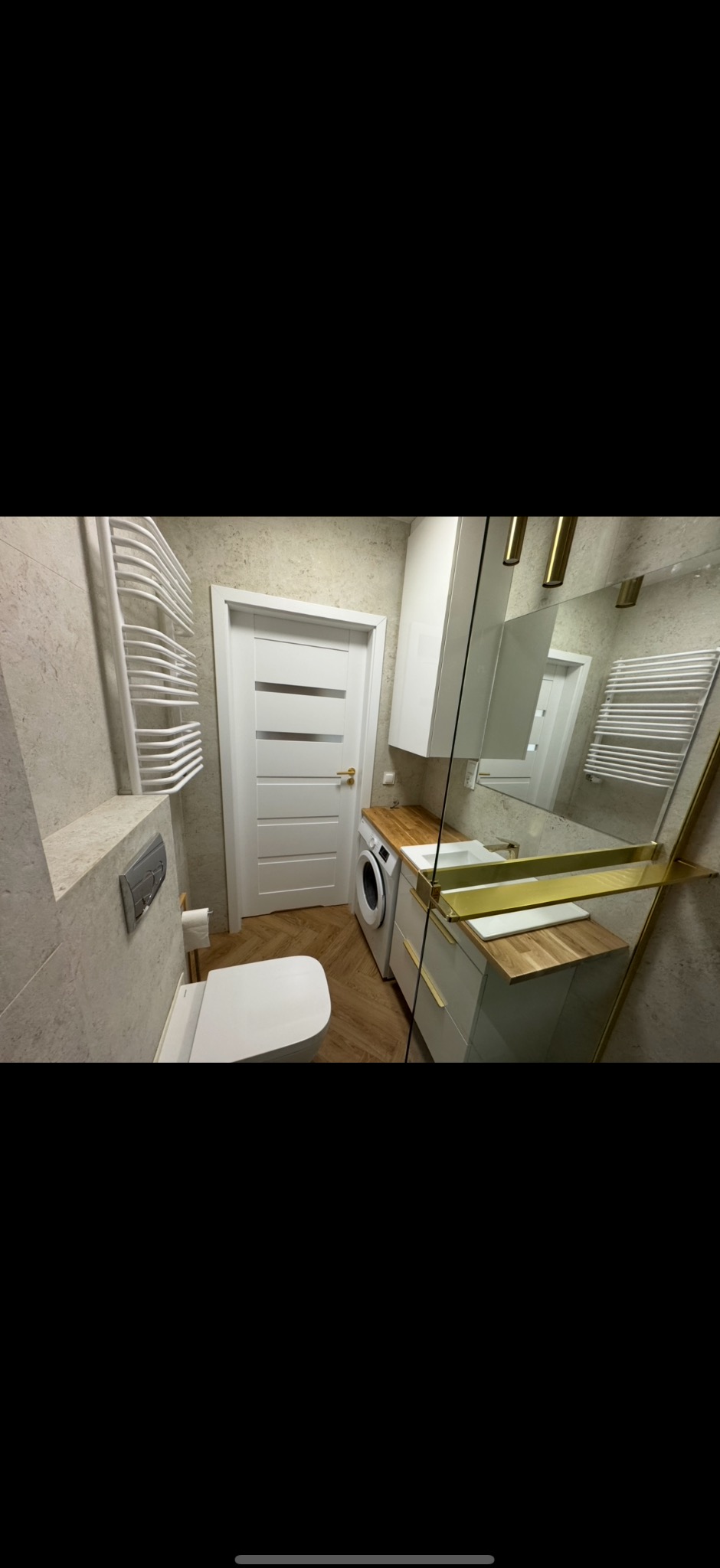 Ukończona, nowoczesna łazienka z pralką, toaletą, grzejnikiem drabinkowym i szafką z drewnianym blatem. Jasne kolory, parkiet w jodełkę, złote akcenty.