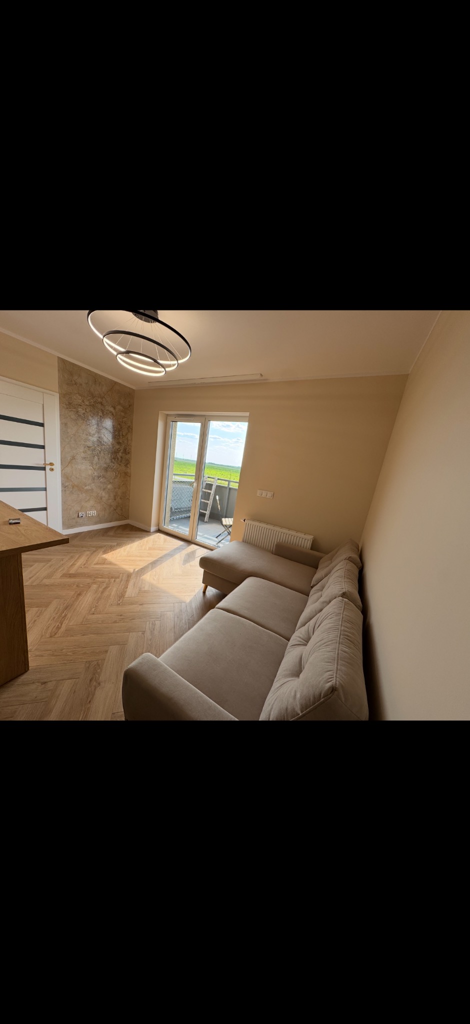 Jasne wnętrze po remoncie: parkiet w jodełkę, beżowa sofa narożna, ściana z efektem marmuru i nowoczesna lampa sufitowa. Wyjście na balkon z widokiem na zieleń.