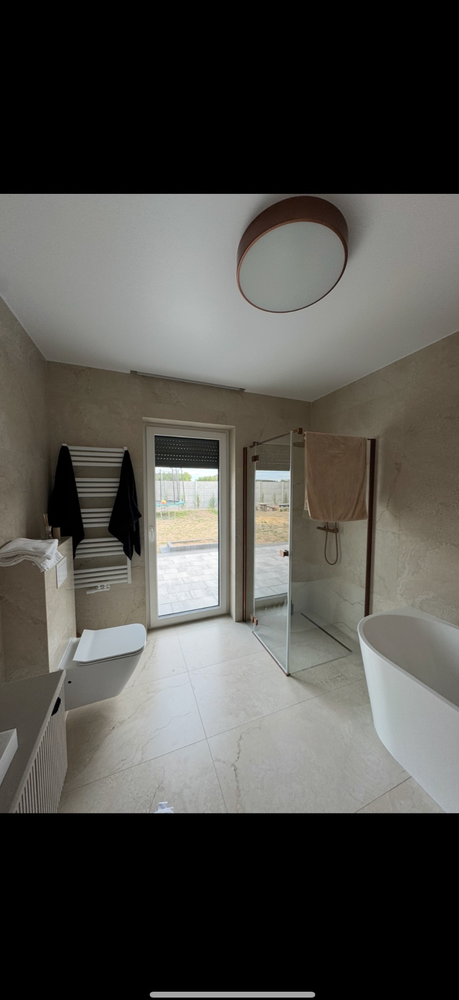 Jasna łazienka z beżowymi płytkami, kabiną prysznicową z miedzianymi profilami, wolnostojącą wanną i oknem na ogród. Nowoczesny design.