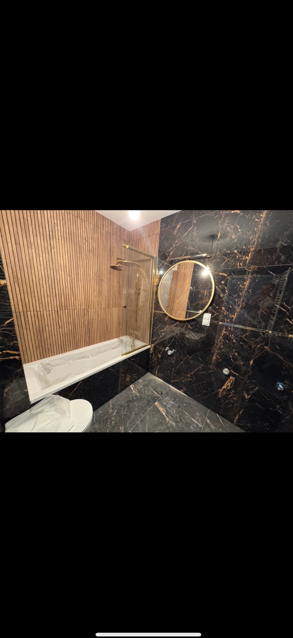 Elegancka łazienka z czarnymi płytkami imitującymi marmur, drewnianą ścianą, okrągłym lustrem w złotej ramie i kabiną prysznicową ze złotymi elementami.