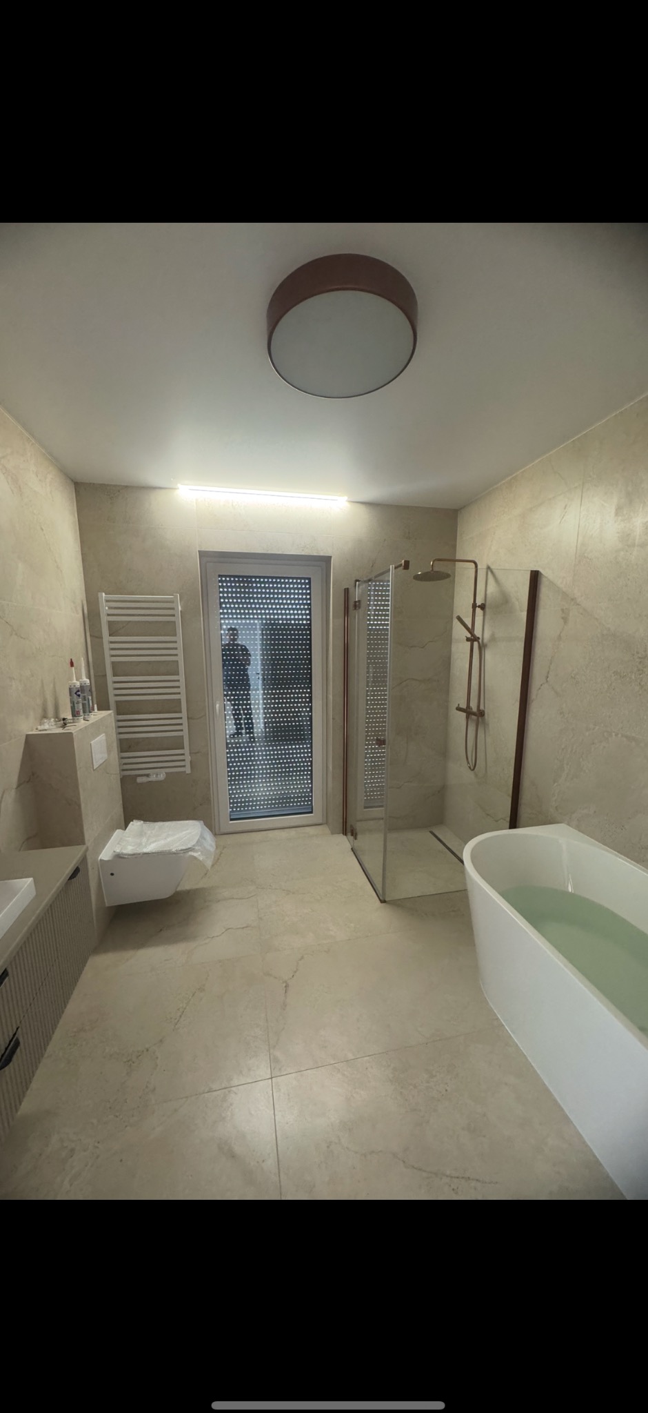 Kompleksowo wykończona łazienka z beżowymi płytkami, kabiną prysznicową z miedzianymi detalami i wolnostojącą wanną wypełnioną wodą. Widoczny grzejnik drabinkowy i podwieszana toaleta.