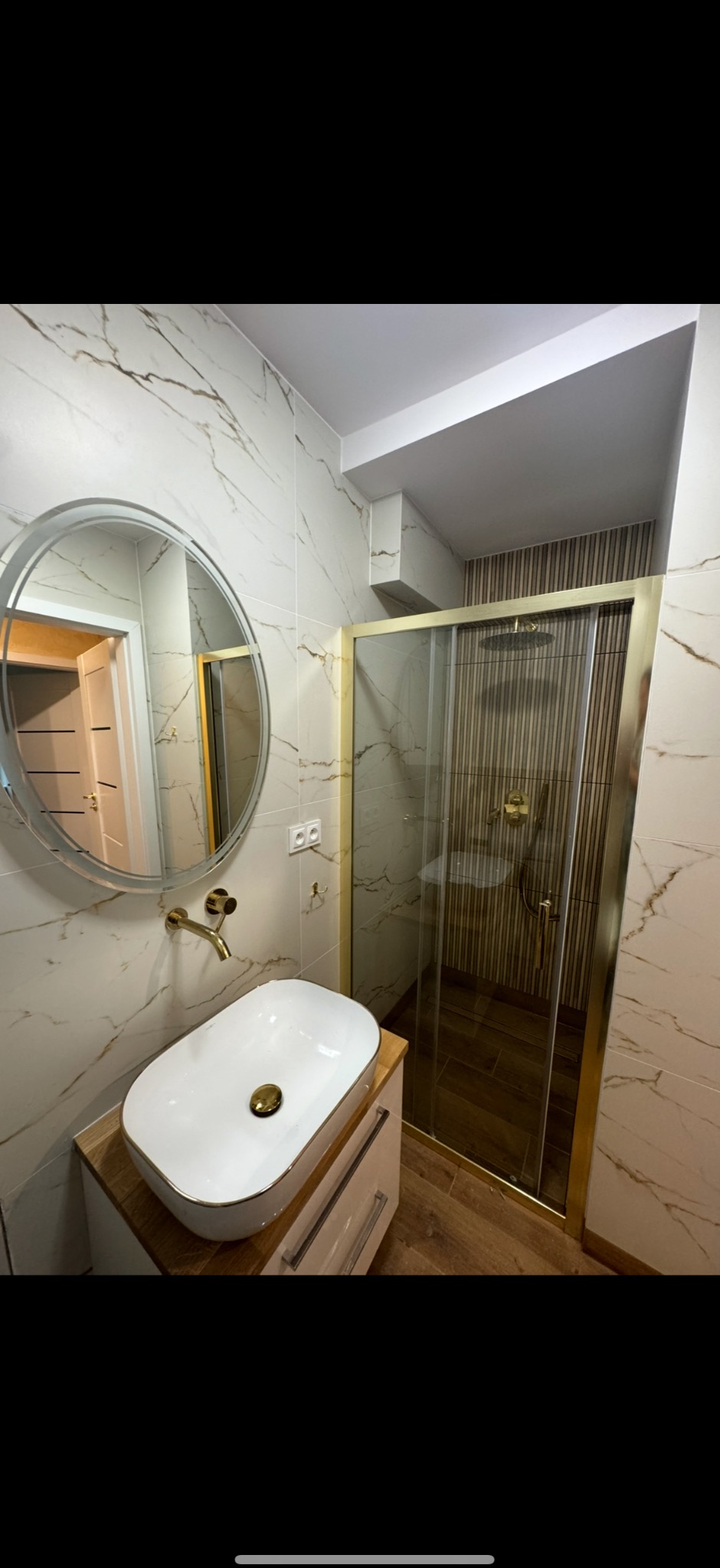 Elegancka łazienka po remoncie: białe płytki z marmurowym wzorem, owalne lustro, umywalka nablatowa i kabina prysznicowa ze złotymi ramami.