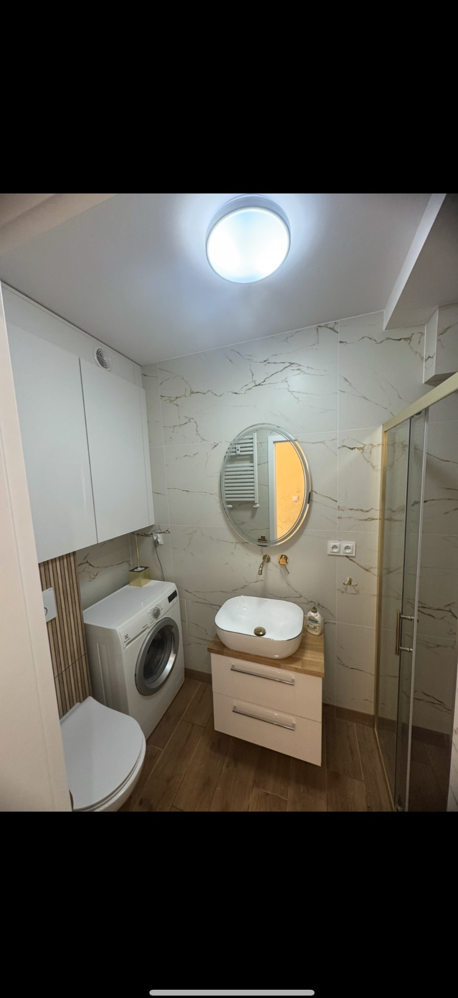 Jasna łazienka po remoncie: marmurowe płytki, okrągłe lustro z podświetleniem, umywalka nablatowa i kabina prysznicowa ze złotymi detalami. Pralka wkomponowana w zabudowę.