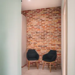Korytarz z ceglaną ścianą, dwoma czarnymi krzesłami na drewnianych nogach i beżową podłogą z płytek. Widoczne oświetlenie szynowe i fragment ściany w kolorze miętowym.